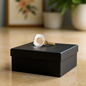 May include: A clear glass cabinet knob with a gold-colored stem rests on a black box. The knob has a circular shape with a visible interior design. The box is rectangular and has a matte finish. The background is blurred.