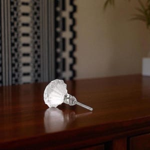 May include: A clear, faceted glass cabinet knob with a silver-toned metal base and screw. The knob is lying on a dark brown wooden surface. The knob's design features a scalloped edge.