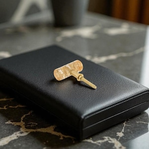 May include: A decorative cabinet knob with a light amber-colored handle and a brass-colored screw. The knob is resting on a black, leather-textured box. The knob's handle is cylindrical and has a marbled appearance.