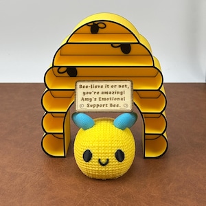 May include: A yellow bee-themed decorative item featuring a crocheted bee and a yellow beehive-shaped structure with black accents. A small sign reads "Bee-lieve it or not, you're amazing!"