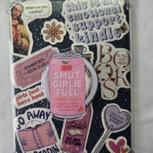 May include: A clear Kindle case decorated with stickers. The stickers include text like "This is my emotional support Kindle," "Smut Girlie Fuel," and "I'm Readin." Other stickers feature books, hearts, and a flower. The case has a glittery background.