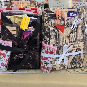 May include: Two fabric book sleeves with front pockets containing pens and bookmarks. The left sleeve features a black dragon print with a black ribbon, while the right sleeve displays a skeleton print with a white ribbon. Both have cherry-patterned accents.
