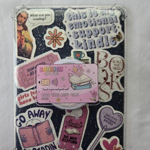 May include: Clear phone case with a dark blue background covered in stickers. Stickers include a pink "Bookish Bank" card, a "Go Away I'm Readin" sticker, and a "This is an emotional support Kindle" sticker. The case has a pop-up grip.