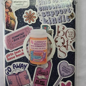 May include: A Kindle case decorated with stickers. The stickers include phrases like "Emotional Support Kindle," "Romantasy Book Club," and "Currently Medicating with Romantasy." The case has a dark blue background with glitter.