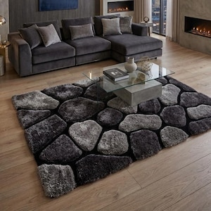May include: A rectangular area rug featuring a textured, multi-tonal grey and charcoal pattern of irregular, stone-like shapes separated by black lines. The rug has a high-pile, shaggy surface and is positioned on a light-coloured wooden floor.