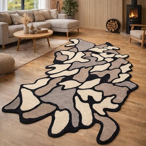 May include: A modern area rug with an abstract design. The rug features a pattern of irregular shapes in shades of beige, gray, and brown, outlined with a black border. It is placed on a hardwood floor in a living room setting.