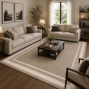 May include: A large rectangular area rug featuring a central beige field framed by multiple concentric borders in shades of cream, light tan, and dark brown. The rug is positioned on a dark wood floor beneath a wooden coffee table in a living room.
