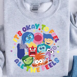 May include: A light gray sweatshirt featuring a colorful graphic of animated characters with the text "It's Okay to Feel All The Feels" in a rainbow circle. The design includes various emotions.