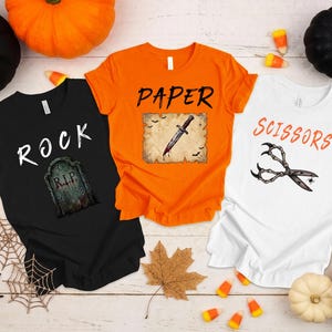 May include: Three short-sleeve t-shirts in black, orange, and white. The black shirt features the text ROCK and a tombstone. The orange shirt features the text PAPER and a dagger graphic. The white shirt features the text SCISSORS and skeleton hands.