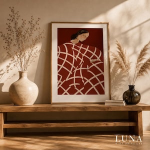 Puede incluir: Una lámina de arte enmarcada que muestra la ilustración estilizada de una persona de pelo oscuro con una prenda de color rojo intenso con un patrón de cuadrícula blanca. La obra está en un marco de madera clara sobre un banco.