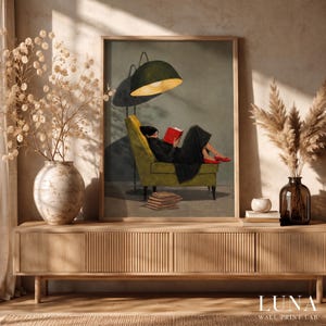 Peut inclure: Une impression d'art encadrée représentant une personne en tenue sombre assise dans un fauteuil vert sous un lampadaire, lisant un livre rouge. Une pile de livres repose au sol. L'impression est exposée sur un buffet en bois avec des plantes séchées.