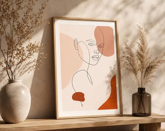 Op de afbeelding: Een ingelijste kunstprint met een zwarte doorlopende lijntekening van een menselijk gezicht tegen een achtergrond van abstracte vormen in tinten lichtroze, perzik en gebrand oranje. De print zit in een lichthouten lijst op een houten ondergrond.