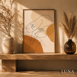 Op de afbeelding: Een ingelijste kunstdruk met een zwarte lijntekening van een persoon met een hoed met brede rand en een hand bij het gezicht, tegen een achtergrond van abstracte beige en terracotta vormen in een rechthoekige houten lijst.