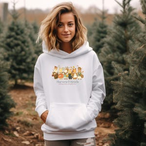 May include: White hoodie with a front pocket and a drawstring hood. The front features a colorful illustration of cartoon animals with the text "Harvest Friends". The design includes various animals in autumn attire.