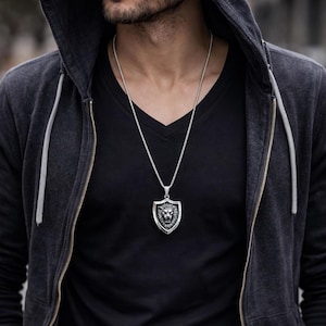 May include: A silver-toned metal pendant necklace featuring a shield-shaped design with a detailed lion head relief in the center. The pendant hangs from a thin, textured silver-toned chain, resting against a black v-neck shirt and a dark hooded jacket.