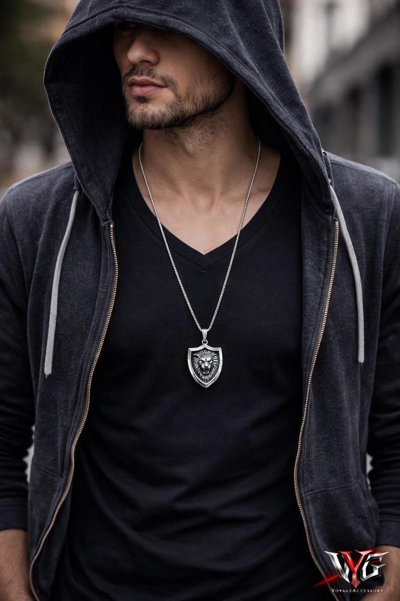 May include: A silver-toned metal pendant necklace featuring a shield-shaped design with a detailed lion head relief in the center. The pendant hangs from a thin, textured silver-toned chain, resting against a black v-neck shirt and a dark hooded jacket.
