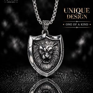 May include: A silver-toned metal pendant necklace featuring a detailed lion head relief centered on a shield-shaped plaque with cracked edge detailing. The pendant hangs from a box chain. Text on the image reads VOYAGE DESIGN, UNIQUE DESIGN, ONE OF A KIND.