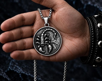 May include: A silver-toned circular pendant featuring a raised scorpion design on a textured background, suspended from a metallic box chain. The pendant displays a polished finish on the scorpion and a dark, recessed surface detail.
