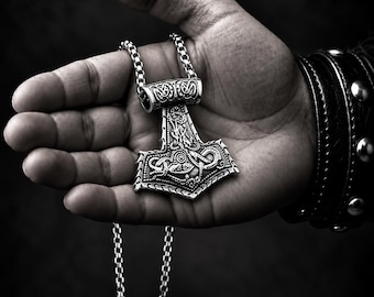 May include: A silver-toned metal pendant shaped like a Mjolnir hammer, featuring intricate embossed knotwork and dragon-like patterns. The pendant is suspended from a matching silver-toned box chain necklace. The text VOYAGE DESIGN is visible.