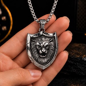 May include: A silver-toned metal pendant shaped like a shield featuring a detailed relief of a lion's head with an open mouth. The pendant has a distressed, weathered finish and hangs from a thick, square-linked silver-toned metal chain necklace.