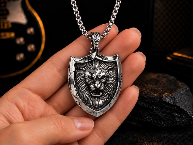 May include: A silver-toned metal pendant shaped like a shield featuring a detailed relief of a lion's head with an open mouth. The pendant has a distressed, weathered finish and hangs from a thick, square-linked silver-toned metal chain necklace.