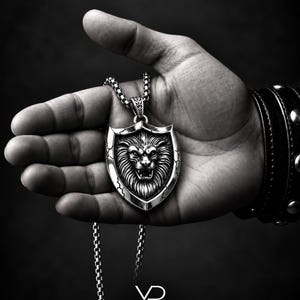 May include: A silver-toned pendant necklace featuring a shield-shaped charm with a detailed lion head relief in the center. The shield has cracked edge detailing and hangs from a metallic box chain. The text VOYAGE DESIGN is visible below.