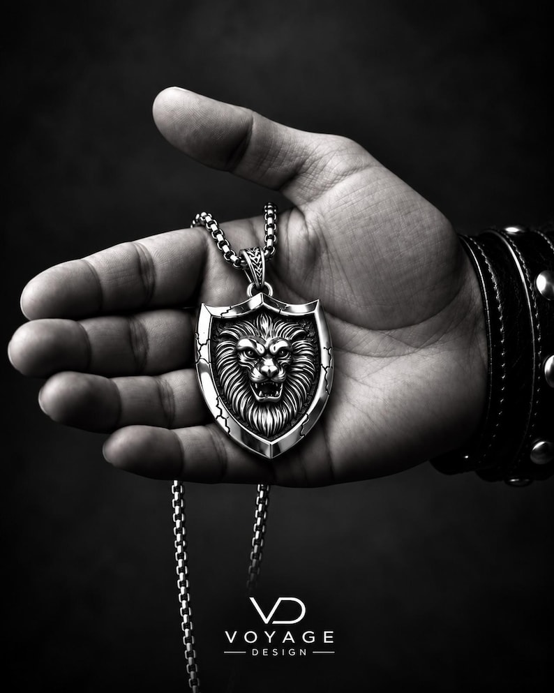 May include: A silver-toned pendant necklace featuring a shield-shaped charm with a detailed lion head relief in the center. The shield has cracked edge detailing and hangs from a metallic box chain. The text VOYAGE DESIGN is visible below.