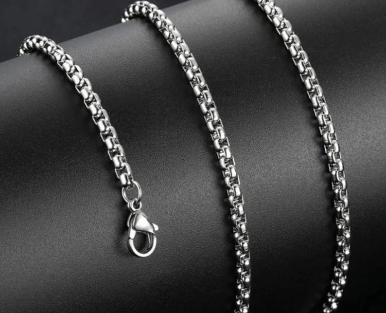 May include: A silver-toned box chain necklace featuring a polished metallic finish and a secure lobster claw clasp. The chain consists of interconnected square links that create a smooth, uniform, and continuous cylindrical appearance throughout.