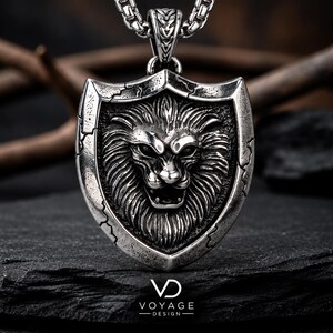 May include: A silver-toned metal pendant shaped like a shield featuring a detailed, three-dimensional lion head relief. The pendant has a weathered, dark finish and hangs from a thick, textured silver chain. The text VOYAGE DESIGN is visible.