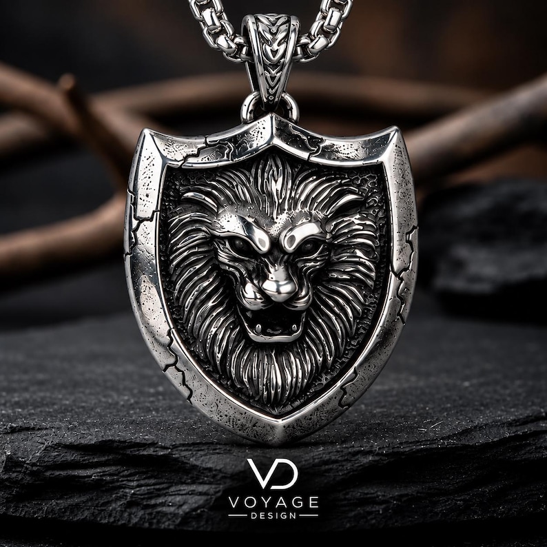May include: A silver-toned metal pendant shaped like a shield featuring a detailed, three-dimensional lion head relief. The pendant has a weathered, dark finish and hangs from a thick, textured silver chain. The text VOYAGE DESIGN is visible.