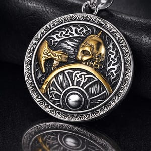 May include: A silver-colored pendant with a Viking design. The round pendant features a gold-colored Viking helmet and hammer, set against a detailed silver background. The design is surrounded by an ornate border. The pendant has a silver loop for a chain.