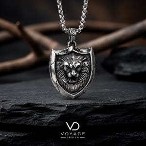 May include: A silver-toned metal pendant necklace featuring a shield-shaped design with a detailed lion head relief in the center. The shield has a cracked texture along the edges and hangs from a thick, textured silver-toned metal chain link.