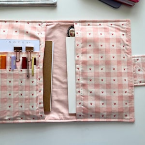 Op de afbeelding: Open roze stoffen organizer met een ruitpatroon en kleine rode hartjes. De organizer is open en onthult vakken met kleurrijke pennen, clips en een notitieblok met het woord "NOTES". Een pen en een notitieboek zijn ook zichtbaar.