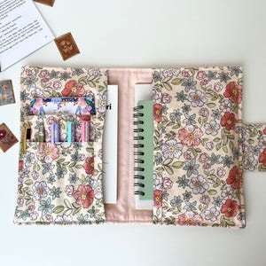 May include: Floral fabric journal cover with multiple pockets, holding pens, cards, and a spiral notebook. The cover features a cream background with pink, red, and blue flowers and green leaves. A button closure secures the cover.