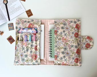 May include: Cream floral journal cover with pockets for pens, cards, and a spiral notebook. The fabric design includes pink, red, and blue flowers with green leaves. A button secures the cover closed.
