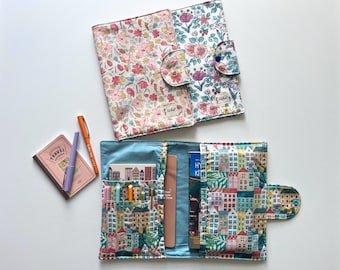 May include: Two fabric journal covers with button closures. One is floral with pink, peach, and white flowers. The other has a colorful cityscape pattern. A small notebook and pens are also visible.