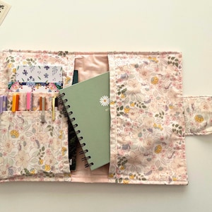 Puede incluir: Funda de cuaderno abierta con estampado floral, con bolsillos para bolígrafos, tarjetas y un cuaderno verde con espiral. La funda es de color rosa claro con un estampado floral en tonos rosas, amarillos y verdes. La funda está abierta, mostrando el contenido.