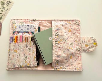 May include: Open floral print notebook cover with multiple pockets holding pens, cards, and a green spiral-bound notebook. The cover is light pink with a delicate floral pattern in shades of pink, yellow, and green. The cover is open, revealing the interior.