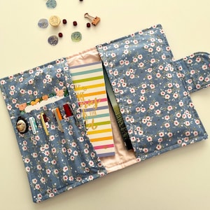 Pode incluir: Uma capa de diário de tecido floral azul com interior rosa, aberta para revelar um caderno listrado colorido. A capa tem bolsos que contêm canetas e um pequeno objeto decorativo. Um fecho de botão é visível no lado direito.