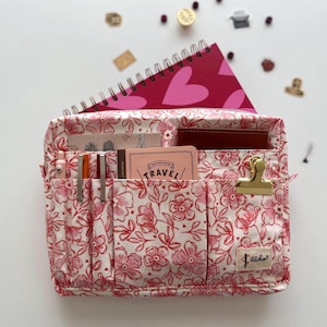 Pode incluir: Uma bolsa organizadora com estampa floral com vários bolsos, contendo um caderno rosa com desenhos de corações, cartões e uma carteira marrom. A bolsa apresenta um clipe dourado e é decorada com um padrão floral vermelho e branco.