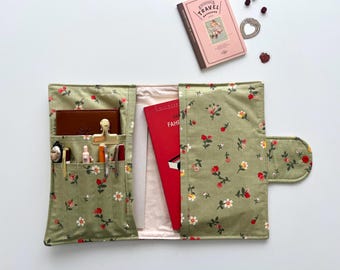 May include: Open green floral fabric notebook cover with pockets, holding a brown leather wallet, pens, and a red book. A pink travel notebook and decorative items are also visible.