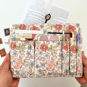 Peut inclure: Un organisateur à imprimé floral avec plusieurs poches et compartiments. Le tissu présente un fond crème avec des fleurs roses, rouges et bleues et des feuilles vertes. L'organisateur est rempli de stylos, de papier et de petits accessoires. Le texte "SINEMSTITCHES" est visible.