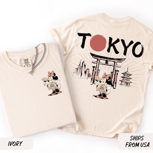 K&ouml;nnte beinhalten: Zwei elfenbeinfarbene T-Shirts. Eines zeigt eine Cartoon-Maus in einem Kimono mit einem F&auml;cher. Das andere zeigt "TOKYO" mit einem roten Kreis, einem japanischen Tor und einer Pagode.