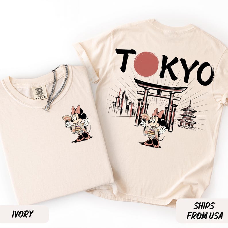 K&ouml;nnte beinhalten: Zwei elfenbeinfarbene T-Shirts. Eines zeigt eine Cartoon-Maus in einem Kimono mit einem F&auml;cher. Das andere zeigt "TOKYO" mit einem roten Kreis, einem japanischen Tor und einer Pagode.