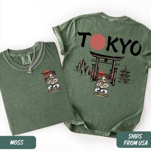 K&ouml;nnte beinhalten: Zwei moosgr&uuml;ne T-Shirts mit einer Zeichentrickfigur in Karate-Uniform. Ein Shirt hat die Figur auf der Vorderseite und das andere das Wort "TOKYO" mit einem roten Kreis und japanischer Architektur. Eine silberne Halskette befindet sich auf einem Shirt.