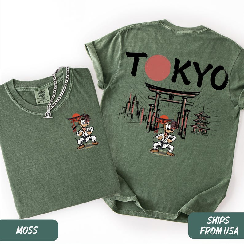 K&ouml;nnte beinhalten: Zwei moosgr&uuml;ne T-Shirts mit einer Zeichentrickfigur in Karate-Uniform. Ein Shirt hat die Figur auf der Vorderseite und das andere das Wort "TOKYO" mit einem roten Kreis und japanischer Architektur. Eine silberne Halskette befindet sich auf einem Shirt.