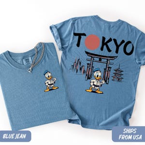 K&ouml;nnte beinhalten: Zwei T-Shirts in Jeansblau. Eines hat eine kleine Donald Duck-Grafik auf der Vorderseite. Das andere hat eine gr&ouml;&szlig;ere Grafik auf der R&uuml;ckseite mit dem Wort "TOKYO", einem roten Kreis und einer japanisch thematisierten Illustration.