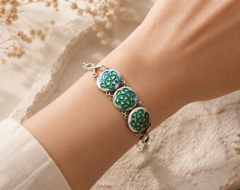 Puede incluir: Pulsera de plata con tres eslabones circulares esmaltados. Cada eslabón presenta un fondo blanco con un diseño de nudo celta azul y verde. La pulsera se lleva en la muñeca.