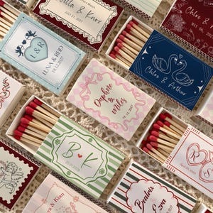 May include: A collection of rectangular matchboxes featuring various custom designs, including names, dates, and illustrations like hearts, swans, and cupids. Each box contains wooden matches with red tips, displayed in a top-down arrangement.