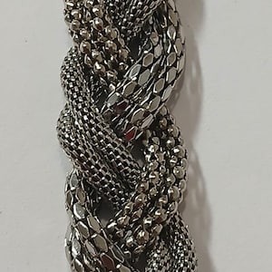 Può includere: Bracciale argentato con un design intrecciato. Il bracciale presenta più trame, tra cui piccole perline, maglie di catena intrecciate e pezzi più grandi, piatti, a forma di scaglia. La chiusura è una barra rettangolare strutturata.